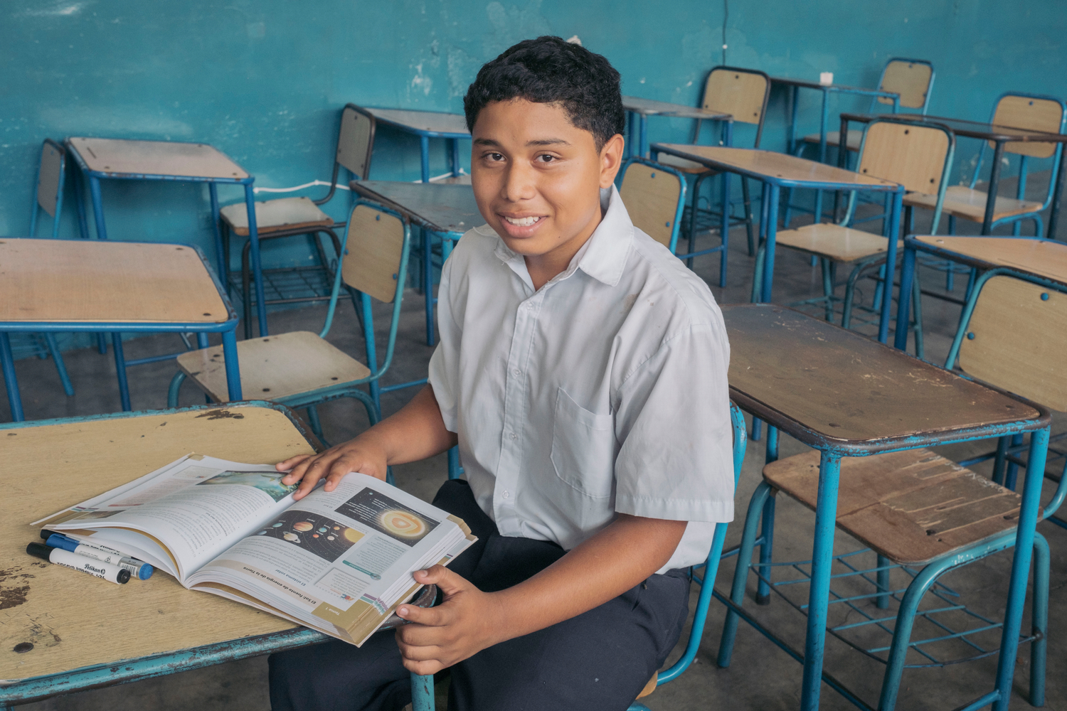 Sentado en un aula, un chico venezolano sonríe a la cámara. El resto de las bancas están vacías.