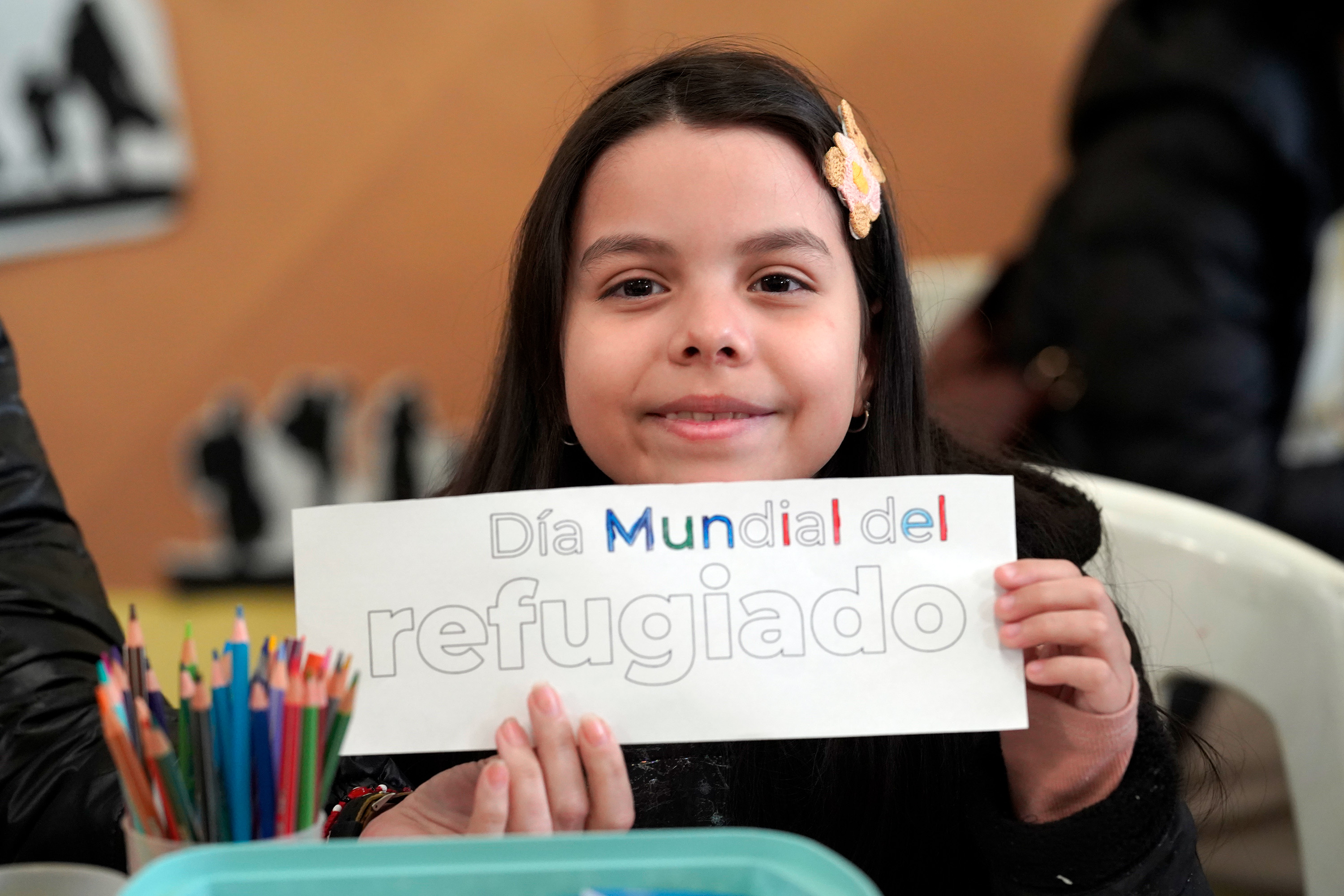 Un niña de cabello largo negro, sonríe mientras sostiene un letrero colorido que dice: Día Mundial del Refugiado