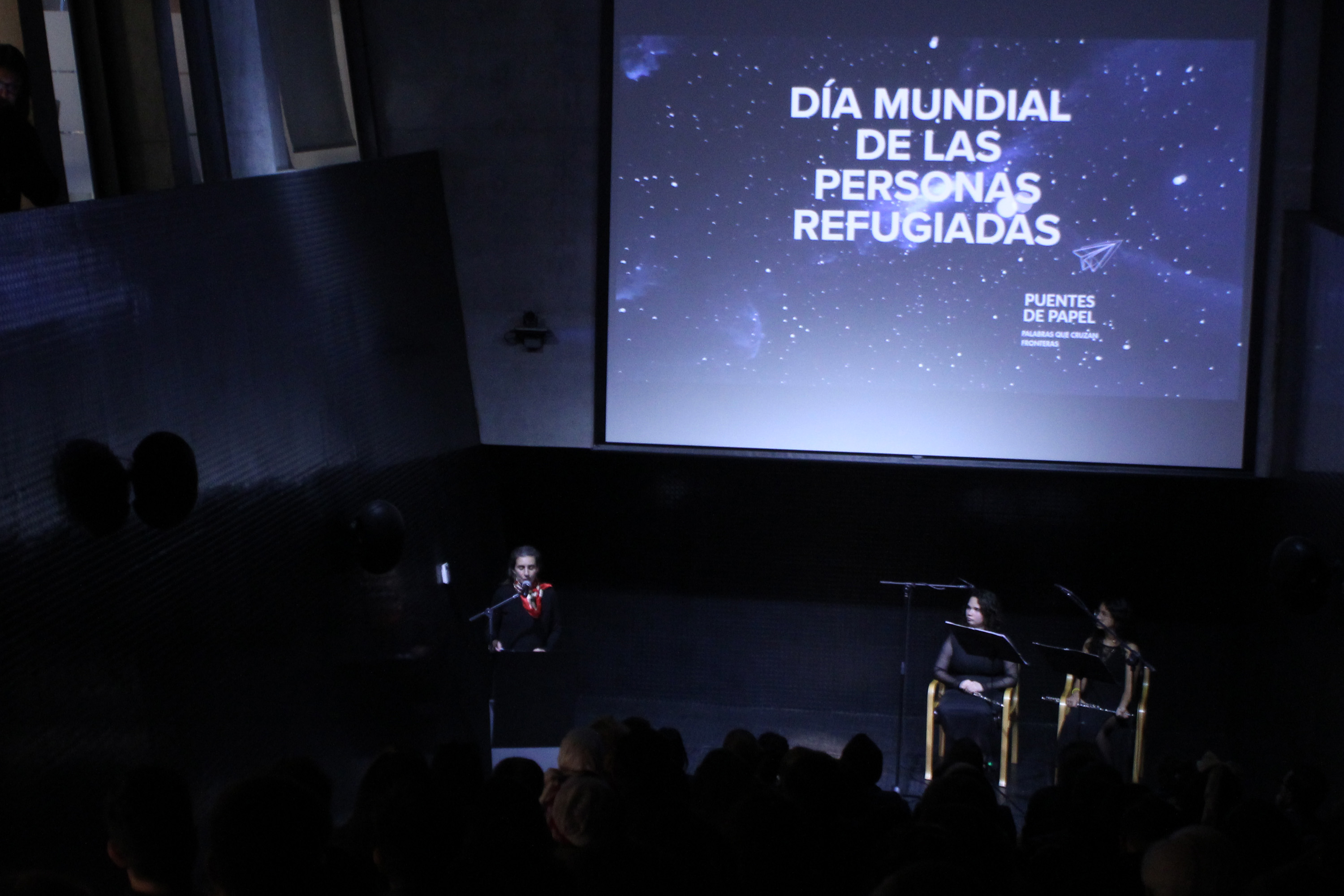 En un auditorio oscuro se lee en una pantalla con letras blancas: DÍA MUNDIAL DE LAS PERSONAS REFUGIADAS