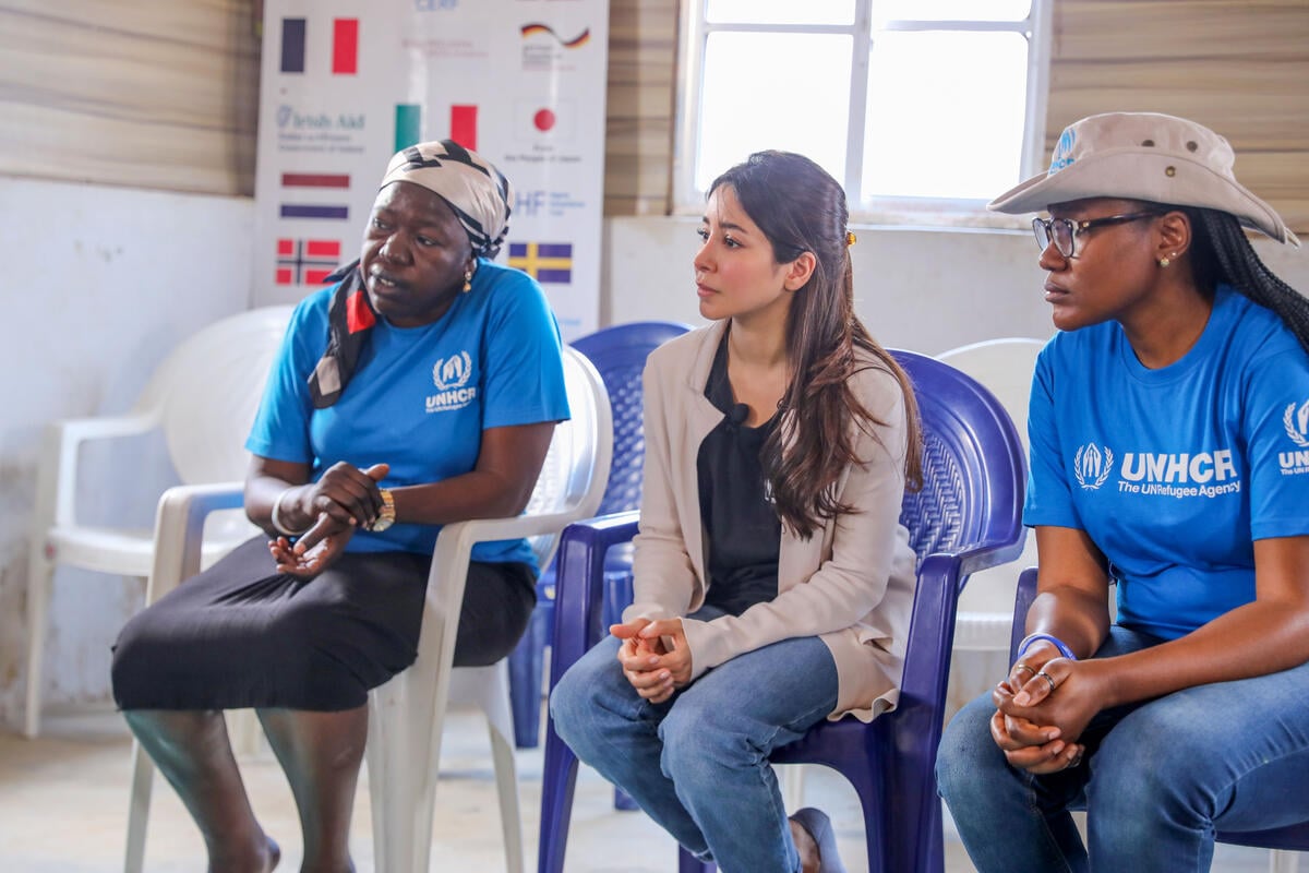 Tres mujeres sentadas una a lado de la otra: en ambos extremos, dos mujeres afro que llevan puestas camisetas azules con el logotipo de ACNUR; al centro, una mujer saudita de cabello largo. Las tres miran y escuchan con atención a alguien fuera de cuadro.