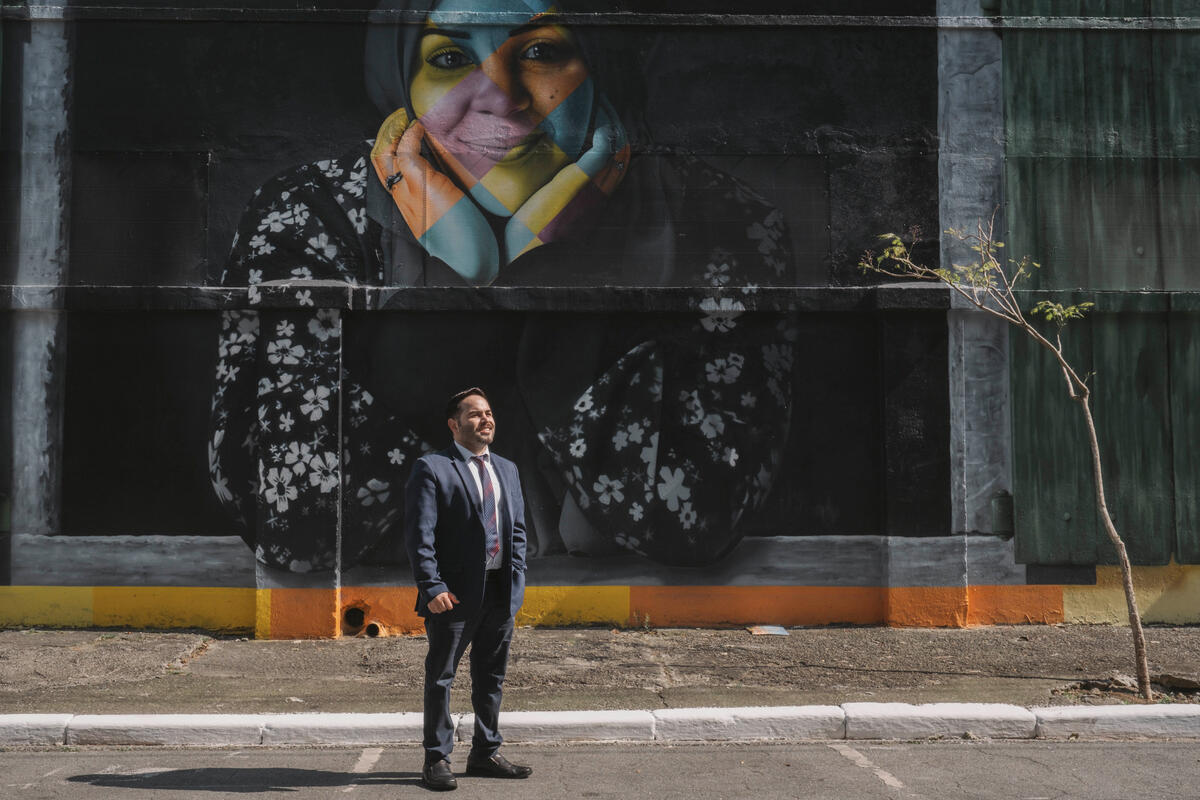 Un hombre vestido con traje y corbata de pie en una calle frente a un muro negro con el rostro de una mujer en colores