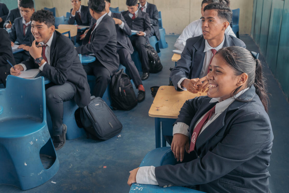 En primer plano una joven sonríe mientras atiende clase, al fondo otros jóvenes en la misma aula