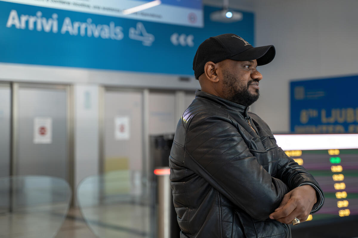 Un hombre vestido con chaqueta de cuero y gorra negras sonríe mirando hacia la lejanía en un aeropuerto. Detrás de él se lee en un letrero «Arrivi/Arrivals» (llegadas)