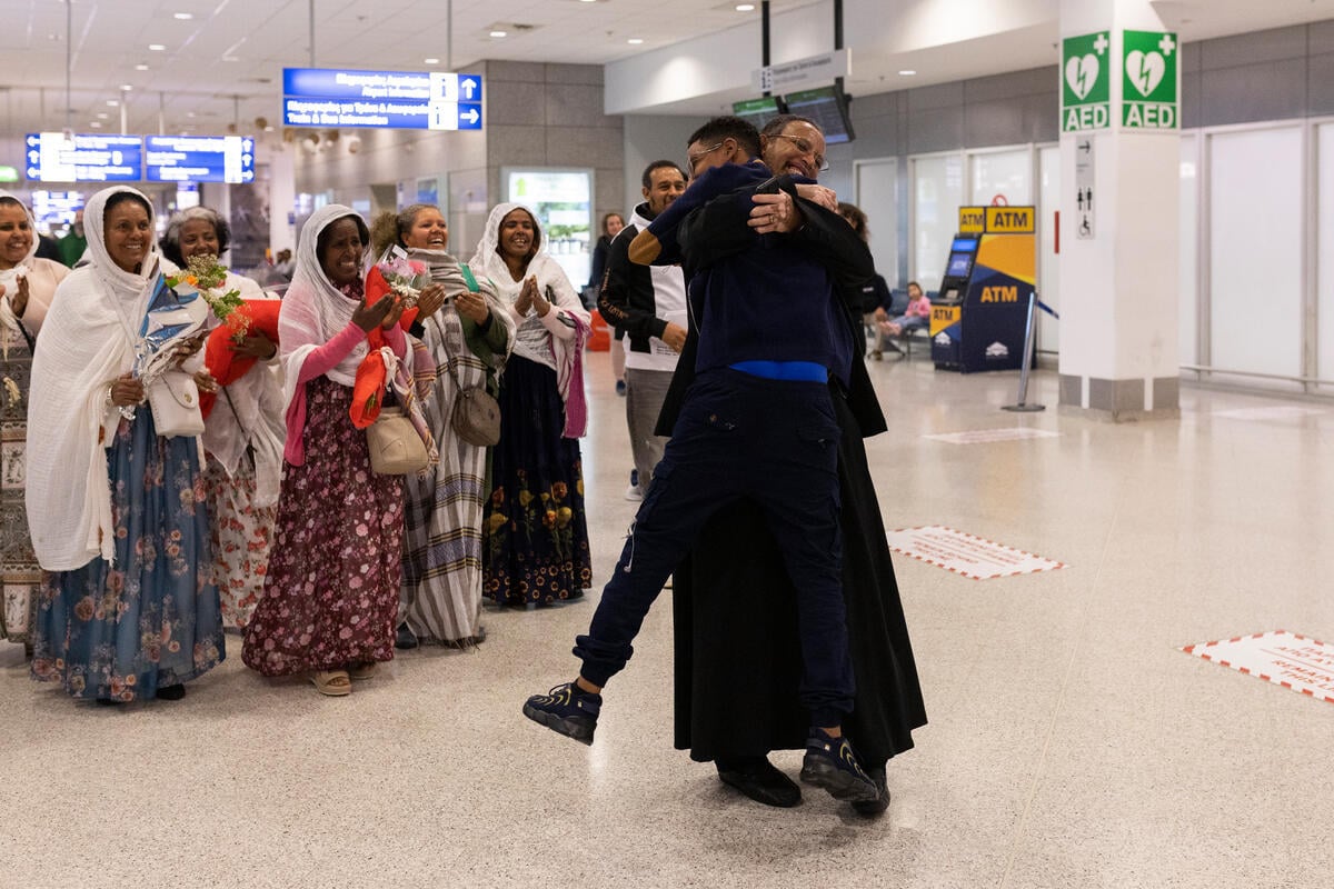Dos hombres vestidos de negro se abrazan fuertemente en un aeropuerto, mientras que un grupo de mujeres permanece cerca, sonriendo y aplaudiendo