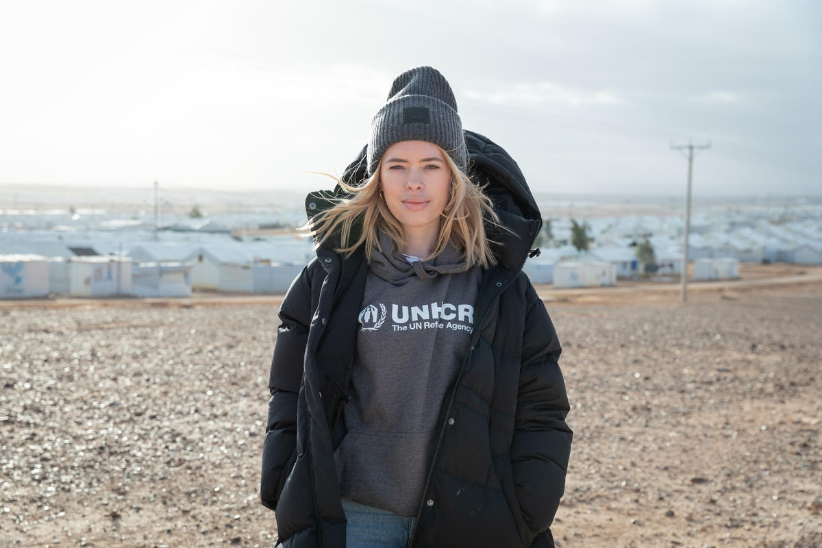 Una mujer caucásica, que lleva puesto un gorro de estambre y una sudadera color gris con el logotipo de ACNUR, sonríe a la cámara; detrás suyo, un campamento de refugiados.