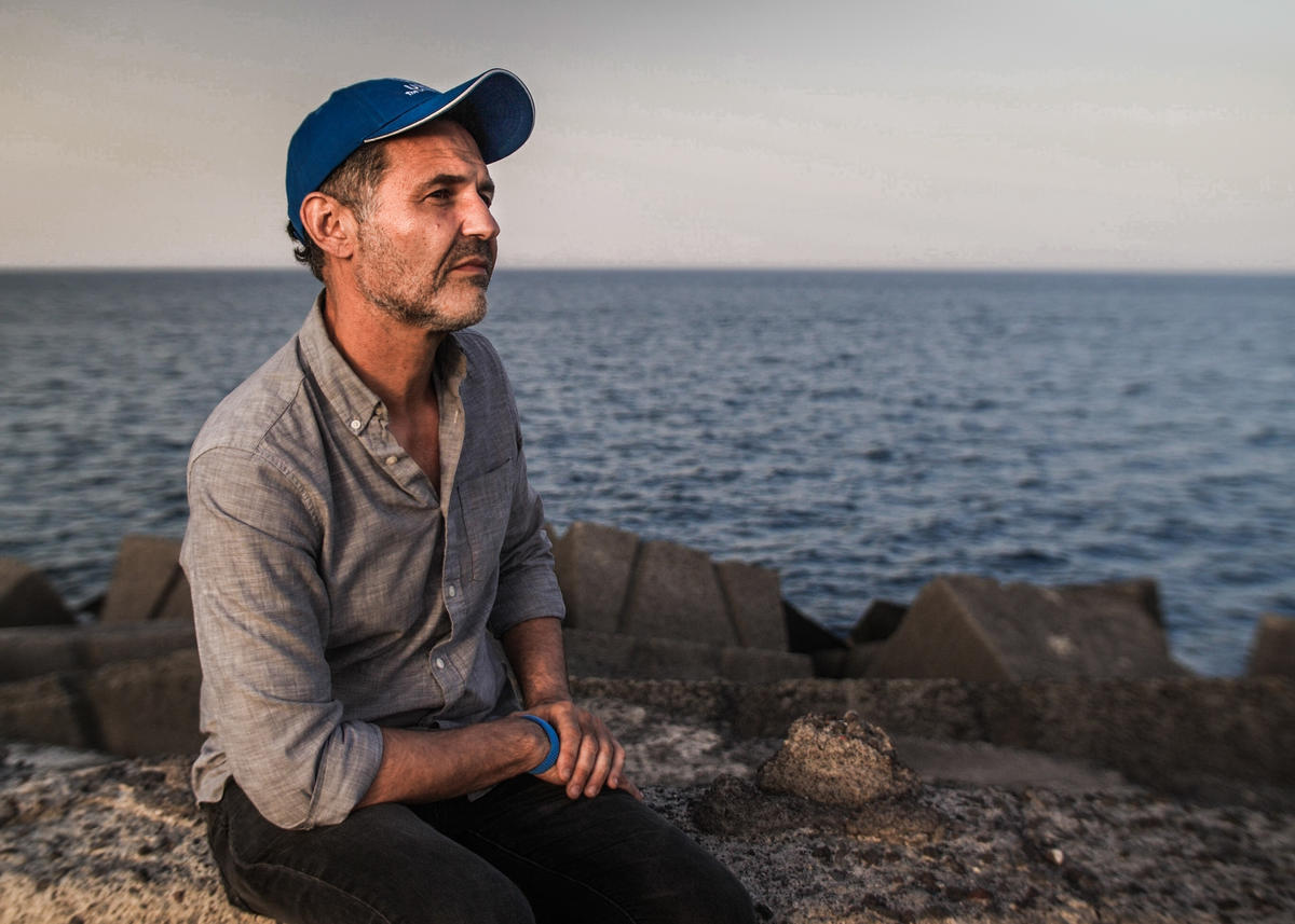 Sentado sobre unas rocas a la orilla del mar, un hombre que lleva puesta una gorra color azul mira al horizonte