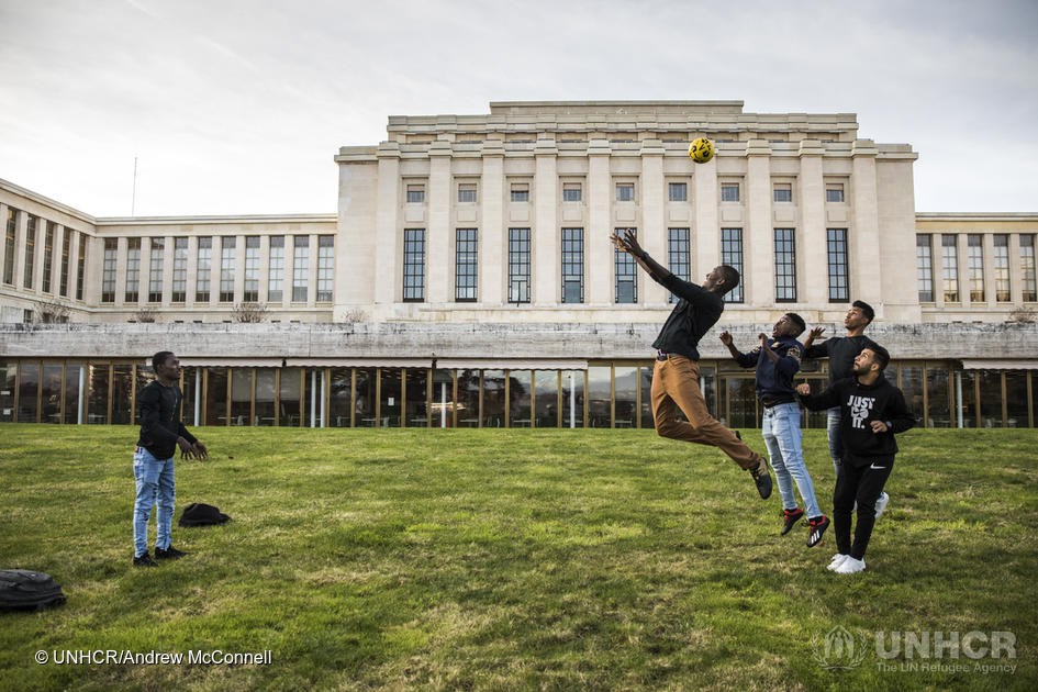 Cuatro hombres juegan al fútbol en un jardín; detrás suyo, un edificio color blanco