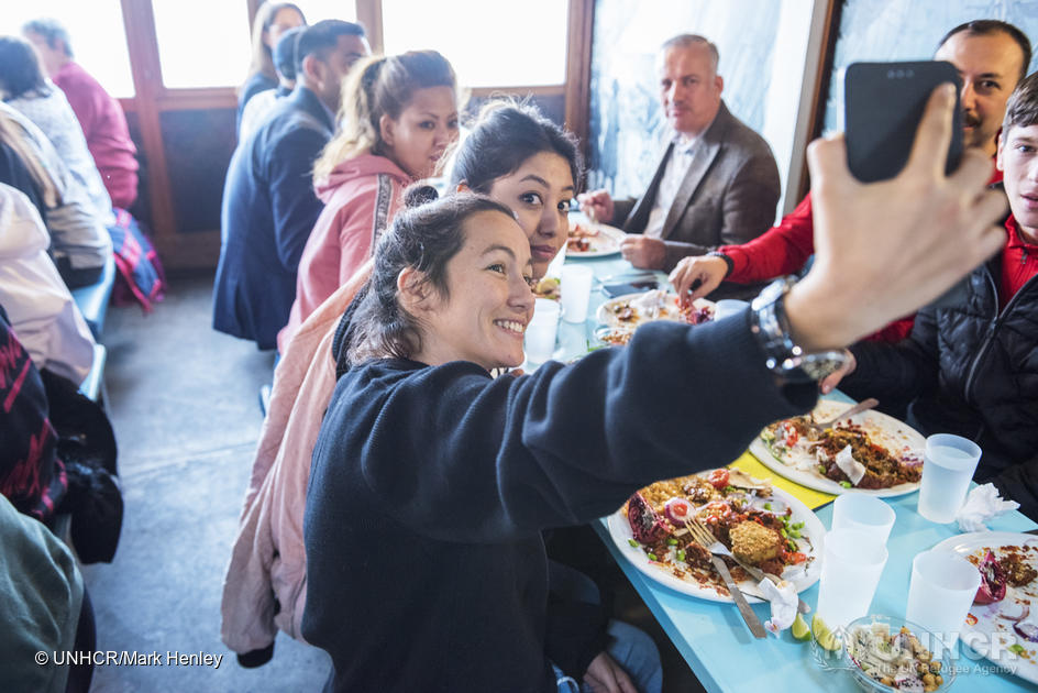Una chica toma una selfi con un grupo de personas que almuerza en una mesa sobre la que hay platos con comida