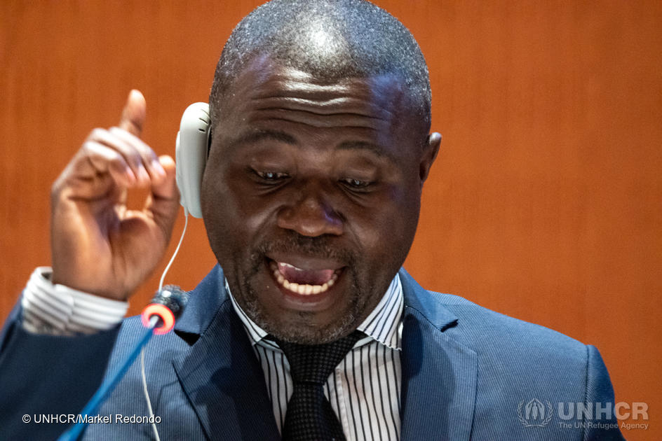 Un hombre ugandés lee un discurso; tiene la mano derecha levantada en un gesto enfático y en el oído derecho lleva dispositivo para escuchar la interpretación