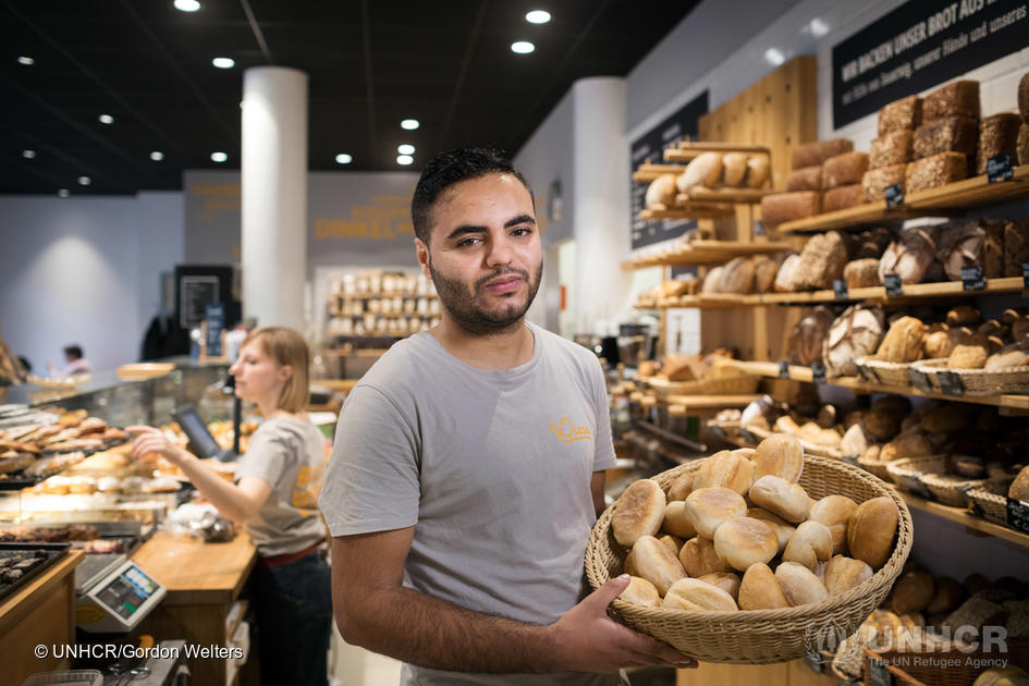 En una panadería, un joven muestra una canasta de pan; detrás suya, una colega atiende a la clientela, que está fuera de cuadro.
