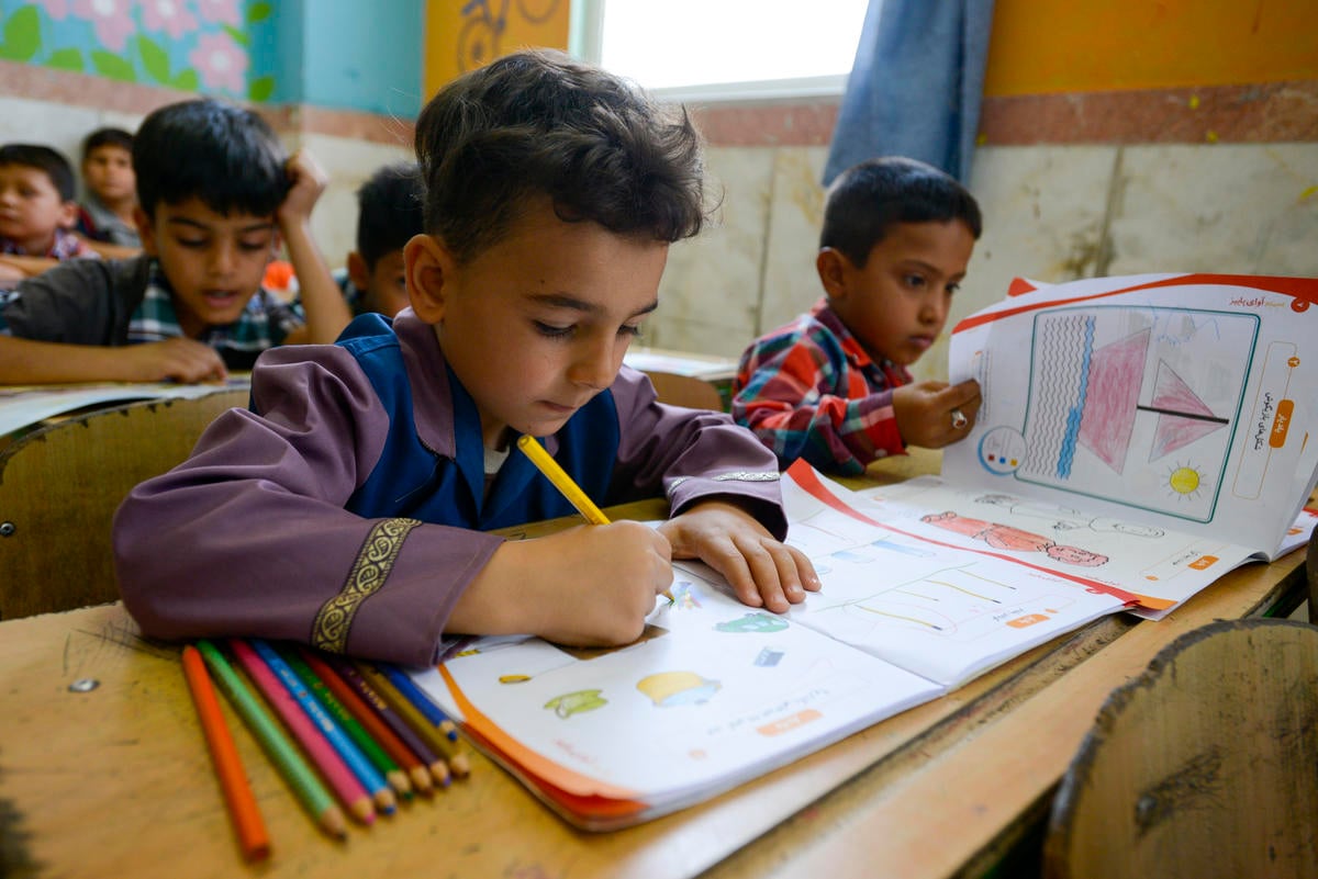 Un niño escribiendo en un cuaderno; a su alrededor, otros niños en un aula