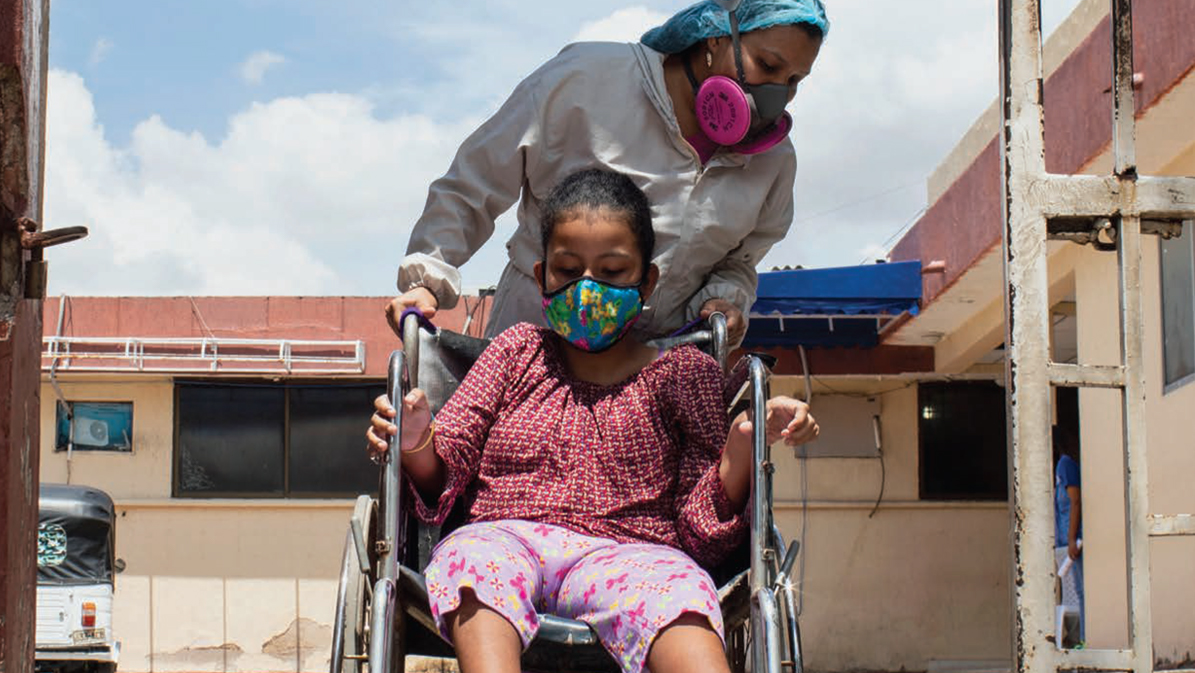 Una mujer ayuda a una niña en silla de ruedas; ambas llevan puesto un cubrebocas. Una mujer ayuda a una niña en silla de ruedas; ambas llevan puesto un cubrebocas.