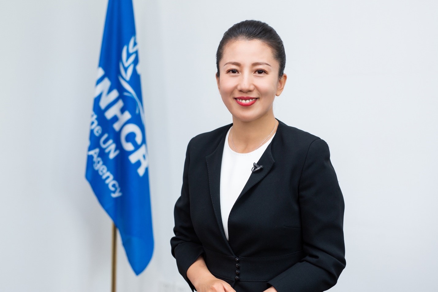 Una mujer china, que lleva puesto un traje color negro, sonríe a la cámara; detrás suyo, una bandera azul con el logotipo de ACNUR en color blanco.