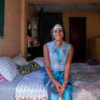 Una mujer joven posa sonriendo sentada al borde de una cama tendida en una habitación colorida.