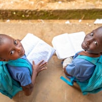 Dos niñas de 7 y 8 años, procedentes de la República Centroafricana, sonríen a la cámara desde donde están sentadas al aire libre, con mochilas a juego en las que se lee 'La educación no puede esperar' y sosteniendo libros abiertos