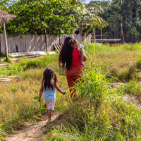 Una mujer camina de espaldas cargando en sus brazos a una niña, mientras otra niña las sigue de cerca.