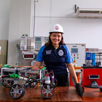 Yuliveth posa para una foto en un laboratorio rodeada dispositivos y mostrando en la mesa delante de ella varios modelos de pequeños robots.