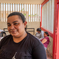 Una mujer posa para la cámara en un salón donde se encuentran varias personas realizando trámites.
