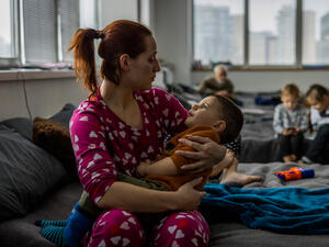 Una mujer sentada en una cama de campamento con su hijo pequeño en brazos en un albergue colectivo para refugiados.