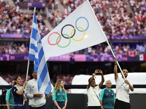 Un hombre porta la bandera olímpica en un estadio abarrotado junto a una mujer que sostiene una bandera griega y otras personas