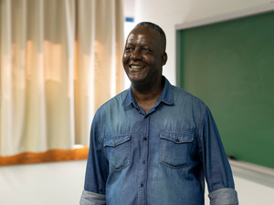Un hombre que viste camisa de mezclilla sonríe en un salón de clases, detrás de él un pizarrón verde y cortinas blancas