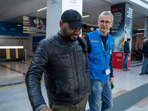 Un hombre de chamarra de cuero negra y gorra negra, camina en un aeropuerto al lado de un funcionaro de ACNUR que viste chaleco azul