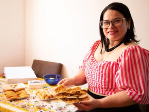 Una mujer que viste blusa a rayas rojas y blancas, y que usa lentes, sonríe para la foto mientras sostiene un plato con arepas venezolanas