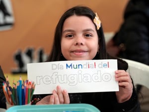 Un niña de cabello largo negro, sonríe mientras sostiene un letrero colorido que dice: Día Mundial del Refugiado