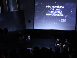En un auditorio oscuro se lee en una pantalla con letras blancas: DÍA MUNDIAL DE LAS PERSONAS REFUGIADAS
