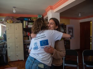 Dos mujeres se abrazan, una de ellas porta una playera blanca con logos de ACNUR y la Unión Europea