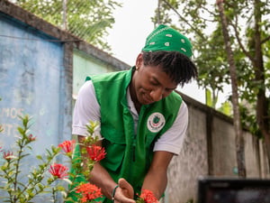 Un joven con un pañuelo verde en la cabeza y chaleco verde, toma entre sus manos unas flores en un jardín