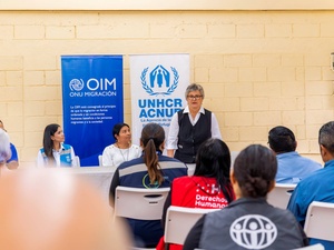 Una mujer hablando desde una mesa de ponencia en un evento frente a un público sentado en una sala, carteles con los logos de ACNUR y OIM están al fondo.
