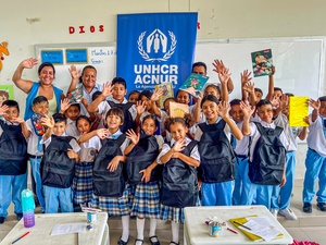 Una veintena de niñas y niños y tres profesores saludan a la cámara en un salón de clases