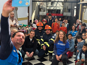 Al menos una decena de niñas y niños posan para la foto junto a ocho adultos, entre ellos dos con vestimenta de ACNUR en color azul y otros dos hombres con uniforme y casco de bombero