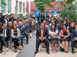 Decenas de personas sentadas en filas en un jardín. Al fondo se observan dos letreros de ACNUR en color azul