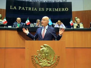 Un hombre abre sus manos mientras habla detrás de un podio, ante una fila de personas que observan desde una plataforma elevada detrás de él