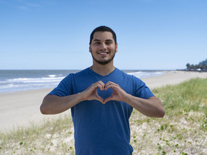 Un joven posa para la cámara en una playa haciendo el gesto del corazón con las manos.