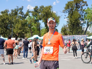 Un hombre con camiseta naranja y shorts negros, porta una medalla colgada al cuello mientras levanta su pulgar izquierdo al sonreir para la foto