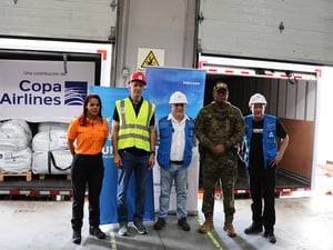 Una mujer que viste pantalon negro y camisa naranja, seguida de 4 hombres que tienen vestimenta de trabajo, tres de ellos usan casco