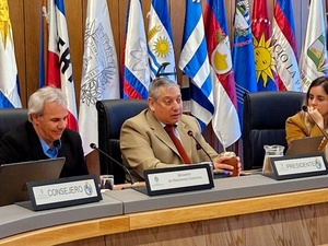 Dos hombres y una mujer sentados en una mesa semicircular con banderas de diferentes países al fondo