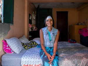 Una mujer joven posa sonriendo sentada al borde de una cama tendida en una habitación colorida.