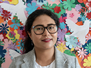 Una mujer con lentes de aros negros, frente a un muro decorado con flores de colores hechas de papel.