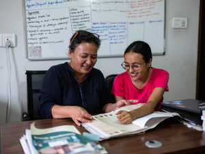 Dos mujeres nepalíes, sentadas frente a un escritorio, revisan un documento; detrás suyo, un pintarrón lleno de texto