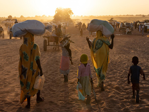 Personas refugiadas sudanesas y sus hijos caminan hacia el sol, llevando sus pertenencias sobre la cabeza o en carretas