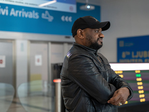 Un hombre vestido con chaqueta de cuero y gorra negras sonríe mirando hacia la lejanía en un aeropuerto. Detrás de él se lee en un letrero «Arrivi/Arrivals» (llegadas)