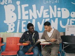 Dos hombres sentados en un salón de clases revisan un libro. Detrás de ellos se lee pintada en la pared la palabra Bienvenidos.