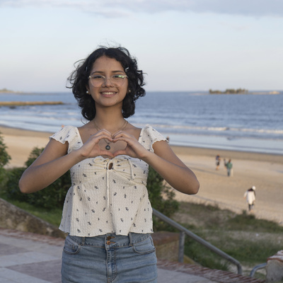 Frente al mar, una joven de cabello corto con anteojos forma un corazón con sus manos