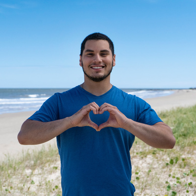 Un hombre joven que viste playera azul y está parado frente al mar, forma un corazón con sus manos