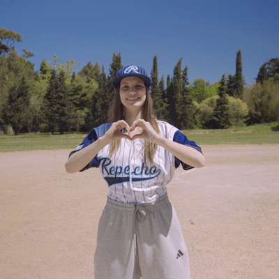Una chica que viste uniforme de un equipo de béisbol, forma un corazón con sus manos en un campo de juego