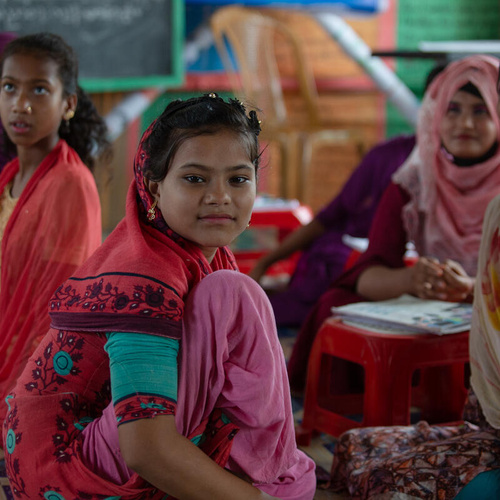 Chicas rohingyas sentadas en el piso. Algunas de ellas miran a alguien o algo fuera de cámara; una de ellas, en el centro de la imagen, ve directo a la cámara.